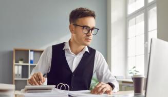 Homem de negócios trabalhando em escritório moderno, focado na tela do computador com documentos e uma caneta na mão, ambiente iluminado e organizado.