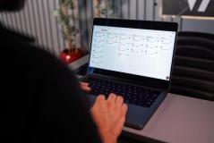 Pessoa de costas operando um notebook aberto em cima de uma mesa cinza de escritório. A tela exibe um software de gestão organizado em colunas verticais com diversas listas de tarefas ou cartões (quadro Kanban). Ao fundo, aparecem uma cadeira de escritório preta e um vaso de planta.