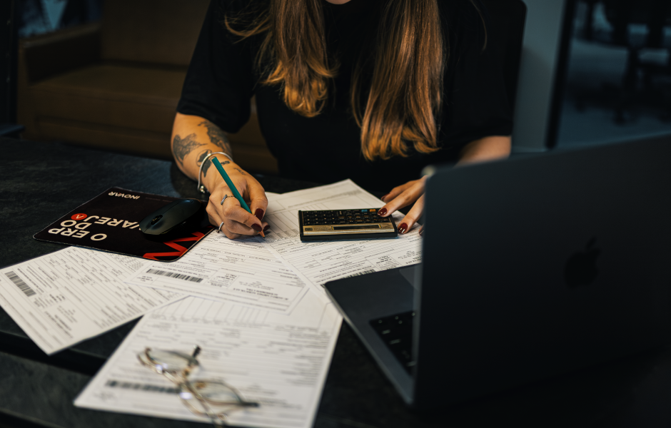 Pessoa sentada à mesa revisando papéis com calculadora, caneta e notebook.