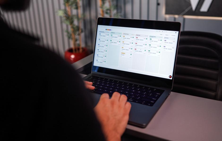 Pessoa de costas operando um notebook aberto em cima de uma mesa cinza de escritório. A tela exibe um software de gestão organizado em colunas verticais com diversas listas de tarefas ou cartões (quadro Kanban). Ao fundo, aparecem uma cadeira de escritório preta e um vaso de planta.