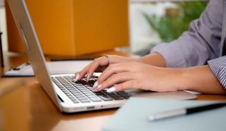 Pessoa digitando em um laptop em um ambiente de escritório, com foco nas mãos e no teclado, incluindo alguns objetos de escritório ao fundo.