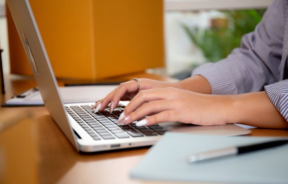 Pessoa digitando em um laptop em um ambiente de escritório, com foco nas mãos e no teclado, incluindo alguns objetos de escritório ao fundo.