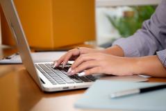 Pessoa digitando em um laptop em um ambiente de escritório, com foco nas mãos e no teclado, incluindo alguns objetos de escritório ao fundo.