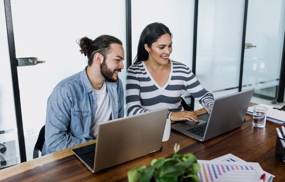Dupla de pessoas trabalhando juntas em um escritório moderno, usando laptops e sorrindo, com ambiente de escritório bem iluminado ao fundo. Ideia de colaboração e produtividade