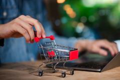 Pessoa usando um laptop em uma mesa de madeira enquanto manipula um carrinho de compras em miniatura, simbolizando compras online ou comércio eletrônico.