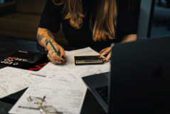 Pessoa sentada à mesa revisando papéis com calculadora, caneta e notebook.