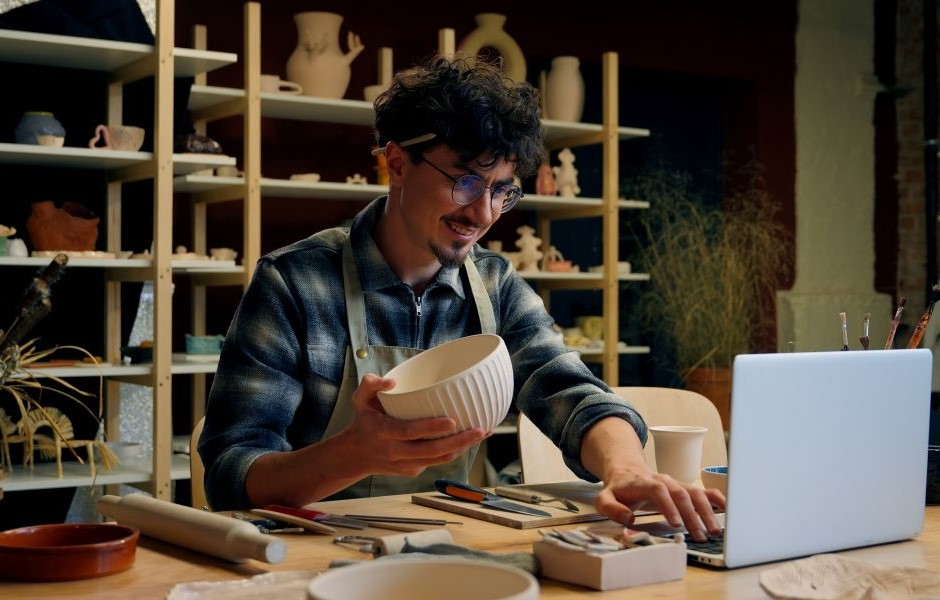 Artesão preparando uma peça de cerâmica em um ateliê, rodeado por várias peças de cerâmica em prateleiras. Pessoa concentrada e criativa.