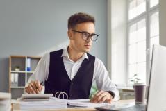 Homem de negócios trabalhando em escritório moderno, focado na tela do computador com documentos e uma caneta na mão, ambiente iluminado e organizado.