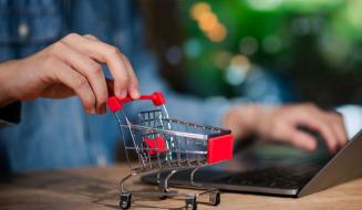 Pessoa usando um laptop em uma mesa de madeira enquanto manipula um carrinho de compras em miniatura, simbolizando compras online ou comércio eletrônico.