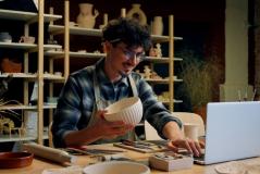Artesão preparando uma peça de cerâmica em um ateliê, rodeado por várias peças de cerâmica em prateleiras. Pessoa concentrada e criativa.