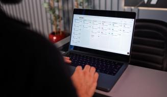Pessoa de costas operando um notebook aberto em cima de uma mesa cinza de escritório. A tela exibe um software de gestão organizado em colunas verticais com diversas listas de tarefas ou cartões (quadro Kanban). Ao fundo, aparecem uma cadeira de escritório preta e um vaso de planta.