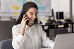 Mulher sorridente trabalhando em escritório moderno, usando computador portátil e telefone celular, com ambiente de trabalho organizado ao fundo.