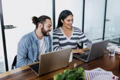 Dupla de pessoas trabalhando juntas em um escritório moderno, usando laptops e sorrindo, com ambiente de escritório bem iluminado ao fundo. Ideia de colaboração e produtividade