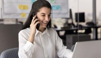 Mulher sorridente trabalhando em escritório moderno, usando computador portátil e telefone celular, com ambiente de trabalho organizado ao fundo.