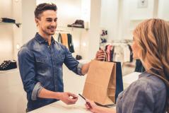 Atendente de loja sorridente entregando saco de compras a cliente. A imagem retrata uma interação simpática no ambiente de moda.