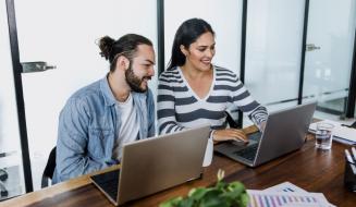 Dupla de pessoas trabalhando juntas em um escritório moderno, usando laptops e sorrindo, com ambiente de escritório bem iluminado ao fundo. Ideia de colaboração e produtividade