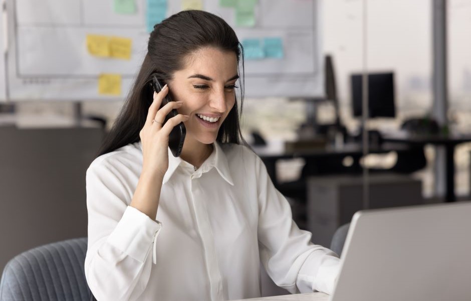 Mulher sorridente trabalhando em escritório moderno, usando computador portátil e telefone celular, com ambiente de trabalho organizado ao fundo.